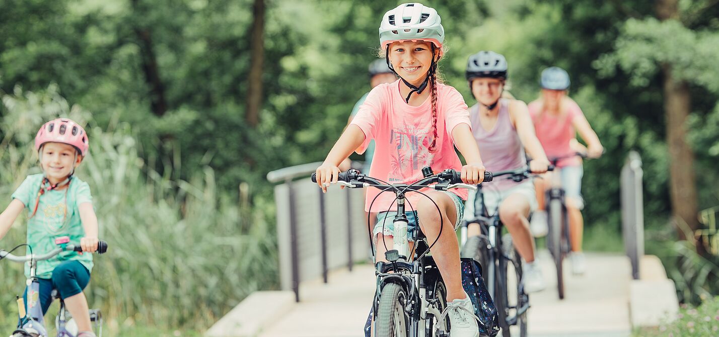 Im Vordergrund fahren freudestrahlende Kinder Fahrrad, im Hintergrund sind die Eltern und eine Brücke verschwommen sichtbar