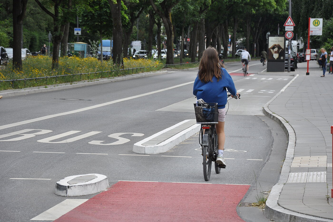 Radfahrerin fährt auf dem Radfahrstreifen. Der ist nach links durch ca. 40 cm breite flache Inseln abgegrenzt. Im Hintergrund eine Bushaltestelle.