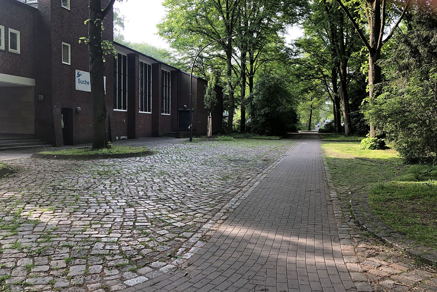 Links eine gepflasterte Fläche, mittig der Radweg durch diese Fläche, rechts Büsche.