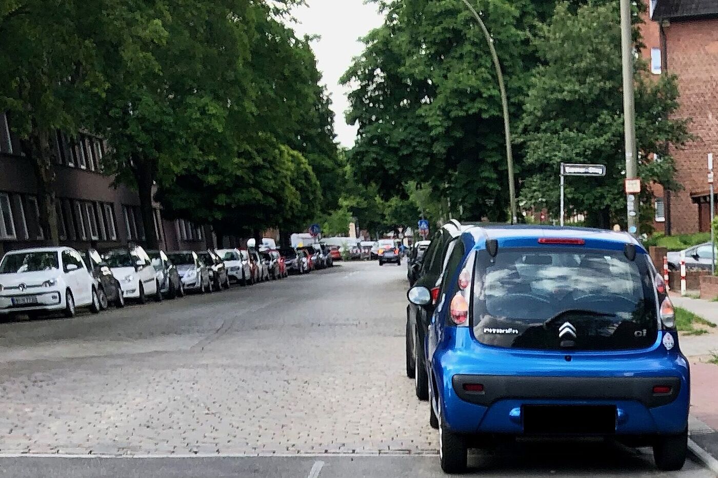 Rechts und links am Fahrbahnrand ist eine durchgehende Reihe Autos geparkt. Dazwischen ist die Fahrbahn aus Kopfsteinpflaster zu sehen.