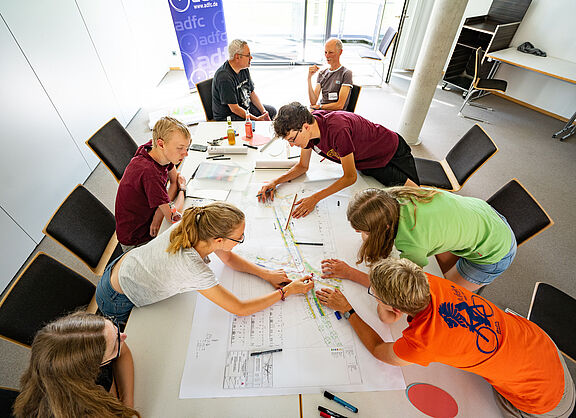 mehrere Personen sind über einem Arbeitstisch gebeugt und bearbeiten gemeinsam Dokumente