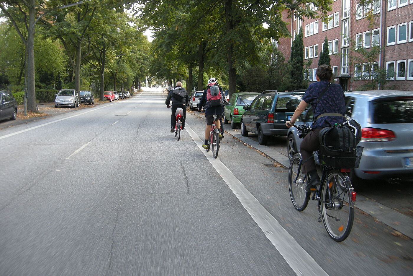 Drei Radfahrende fahren auf bzw. links neben einem Radfahrstreifen. Direkt neben diesem sind Autos geparkt. Es fehlt ein Sicherheitsabstand.