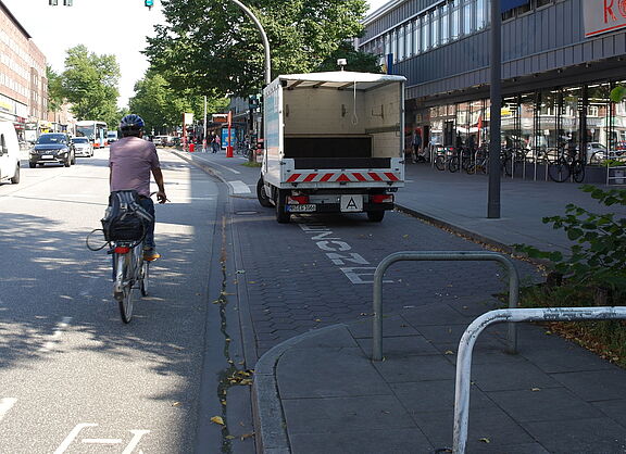 Eine Radfahrerin fährt auf dem Radfahrstreifen. Rechts davon steht auf einer Ladezone ein Fahrzeug der Stadtreinigung. Vor dem Geschäft sind viele Fahrräder an Fahrradbügeln angeschlossen.