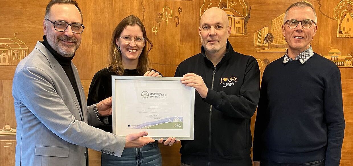 4 Personen im Bild. Florian Mallok übergibt das Zertifikat in einem Bilderrahmen an das Team