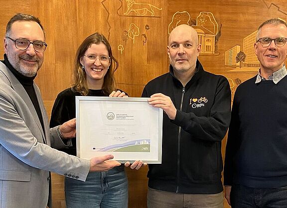 4 Personen im Bild. Florian Mallok übergibt das Zertifikat in einem Bilderrahmen an das Team