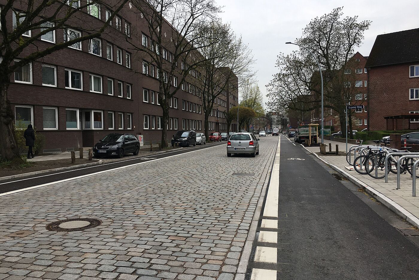 Zwei Fahrstreifen aus Kopfsteinpflaster und je ein asphaltierter Radfahrstreifen daneben. Autos sind in Parkständen abgestellt, Fahrräder stehen an Fahrradbügeln in der Nebenfläche.