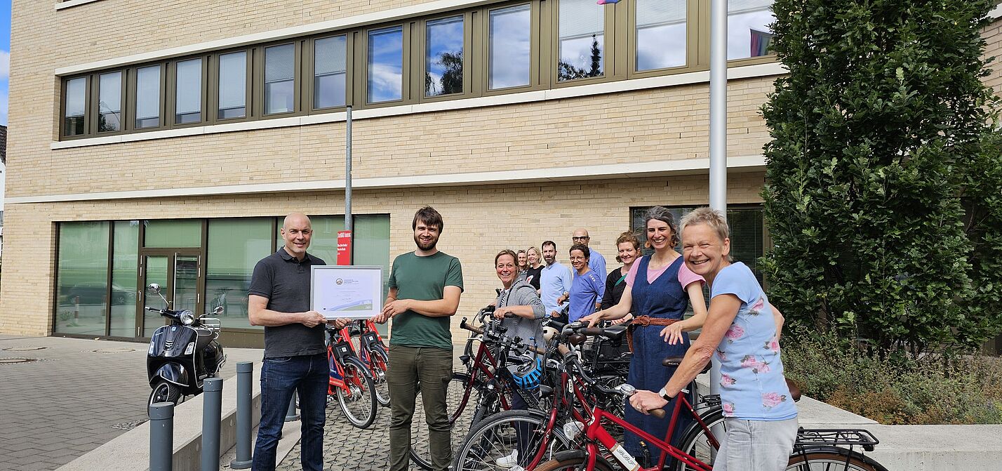 Florian Mallok vom ADFC übergibt ein Zertifikat. Viele fröhlische Menschen mit Rädern stehen dabei und lächeln in die Kamera.