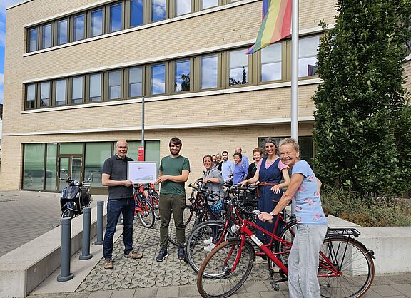 Florian Mallok vom ADFC übergibt ein Zertifikat. Viele fröhlische Menschen mit Rädern stehen dabei und lächeln in die Kamera.