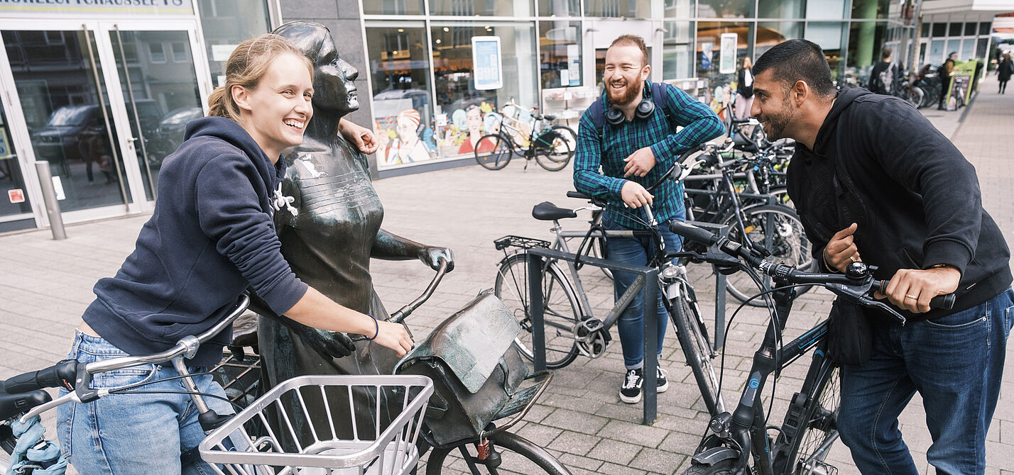 Drei junge Leute fröhlich mit Fahrrädern an einer Radfahrerinnen-Statue