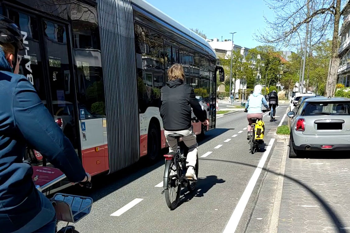 Barcastraße Drei Radfahrende zwischen einem Bus und geparkten Autos auf einem Schutzstreifen